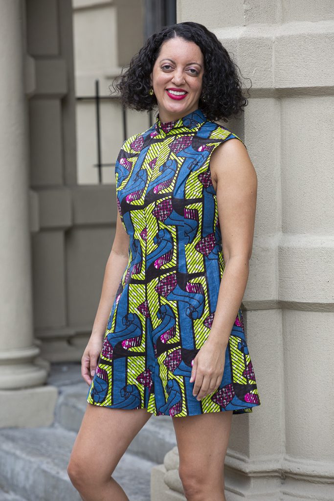 A woman with curly hair smiles while standing outdoors, wearing a sleeveless, patterned dress featuring blue, yellow, and pink designs, in front of a beige building with columns.