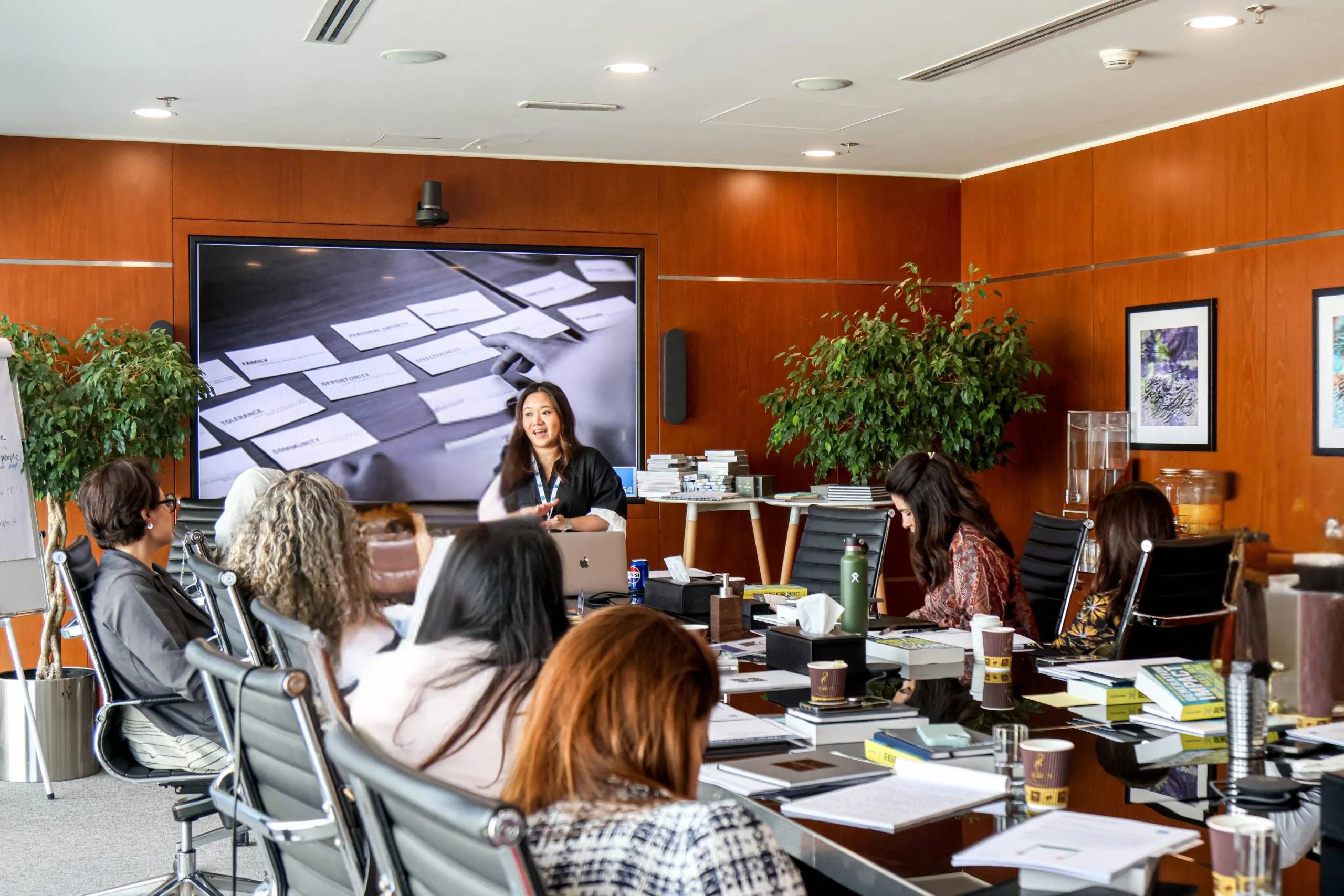 A woman stands at the front of a conference room, presenting to a group of seated people. A large screen behind her displays an image with cards. The room has wooden walls, plants, and office supplies on the table.