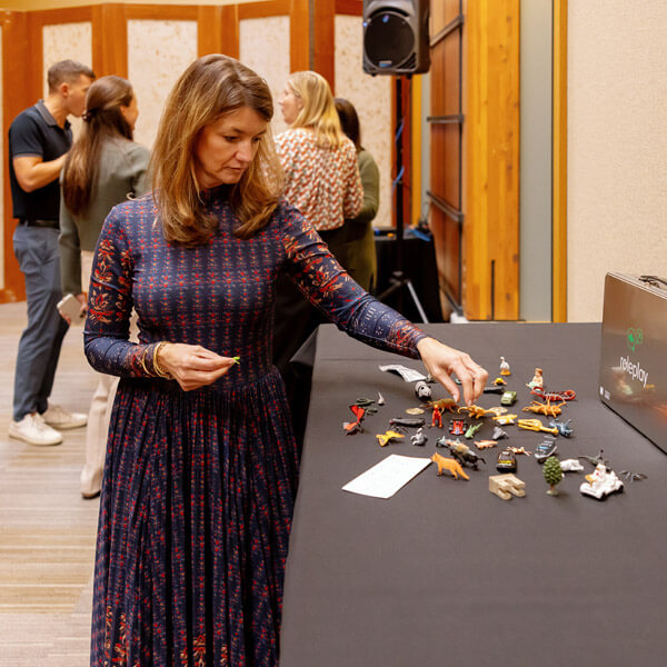 A woman in a patterned dress stands by a table, arranging small animal figurines. Other people are gathered and talking in the background, inside a conference or event space.