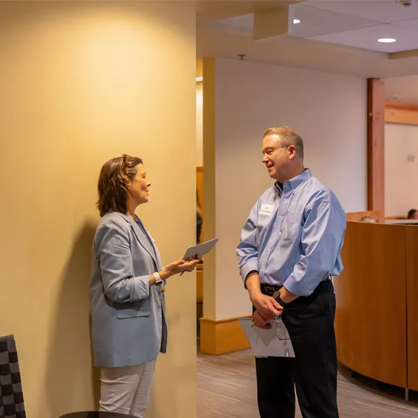 Two adults, a woman and a man, stand and talk in a brightly lit office hallway. Both are dressed in business casual attire, and each holds paperwork while facing each other and conversing.