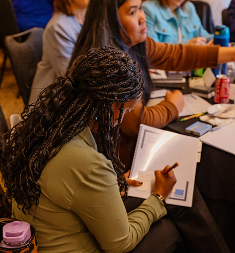 A woman with braided hair writes in a notebook at a table, surrounded by others engaged in a workshop or meeting. Papers, folders, and drinks are scattered on the table.