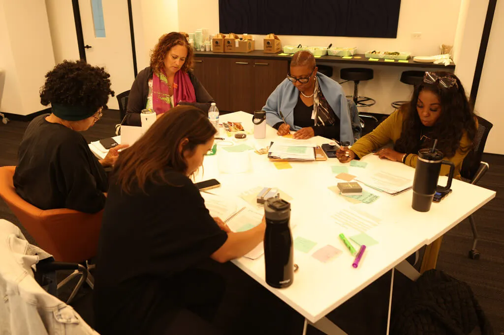 Five women are sitting around a table in an office, focused on reading and writing documents. The table is covered with papers, folders, water bottles, and highlighters. Food trays are visible on a counter in the background.
