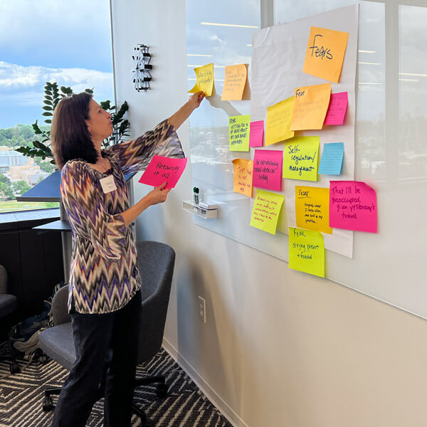 A woman stands by a wall, placing a sticky note among many colorful notes labeled Fears during a brainstorming session in a bright office with large windows.