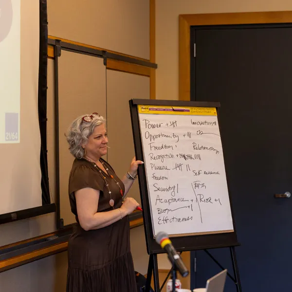 A woman with gray hair stands beside a flip chart covered in notes, pointing to the paper during a presentation in a conference room. A projector screen and a closed door are visible in the background.