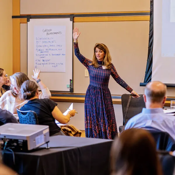 A woman in a long patterned dress stands and raises her hand while speaking to a seated group in a classroom. Several audience members also raise their hands. A whiteboard with notes is visible in the background.