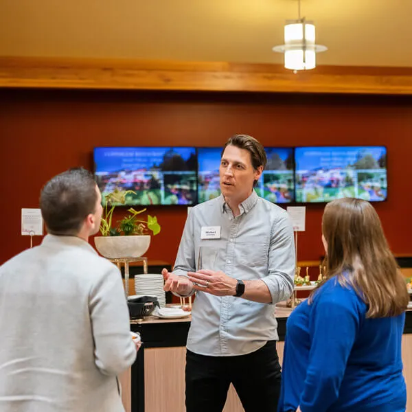 Three people stand talking in a modern indoor space with food and drinks behind them. A man in a light shirt gestures while speaking to two others, with digital screens and a red wall in the background.