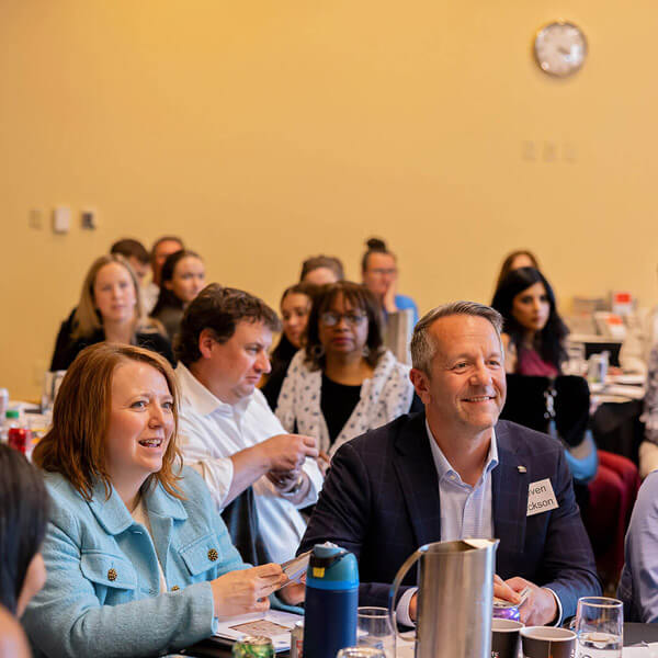 A group of people, including a smiling man and woman in the front, sit at tables in a brightly-lit room, attentively watching a presentation. A clock and various drink containers are visible in the background.