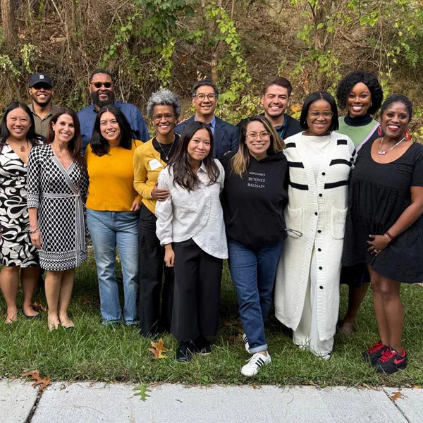 A diverse group of thirteen adults stand together outside on grass, smiling at the camera. Trees and greenery form the background, and everyone is dressed in casual or business-casual clothing.