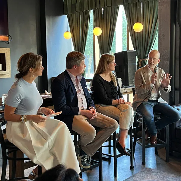 Four people sit on high chairs during a panel discussion in a modern room with green curtains; the man on the far right holds a microphone and gestures as he speaks, while the others listen attentively.