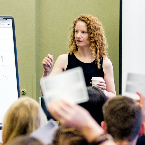 A woman with curly red hair stands near a flipchart, holding a coffee cup and gesturing while speaking to a group of people who are raising papers in a classroom or meeting room.