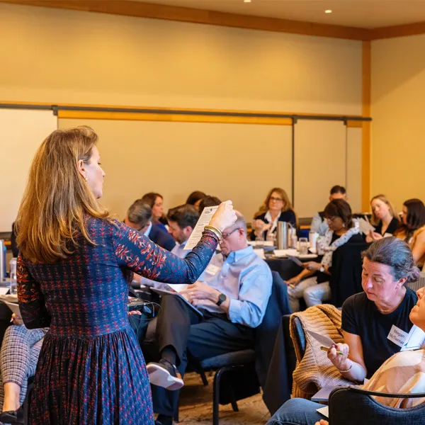 A woman stands at the front of a conference room, holding a paper and addressing a seated group of adults who are listening attentively, some taking notes. A whiteboard is visible in the background.