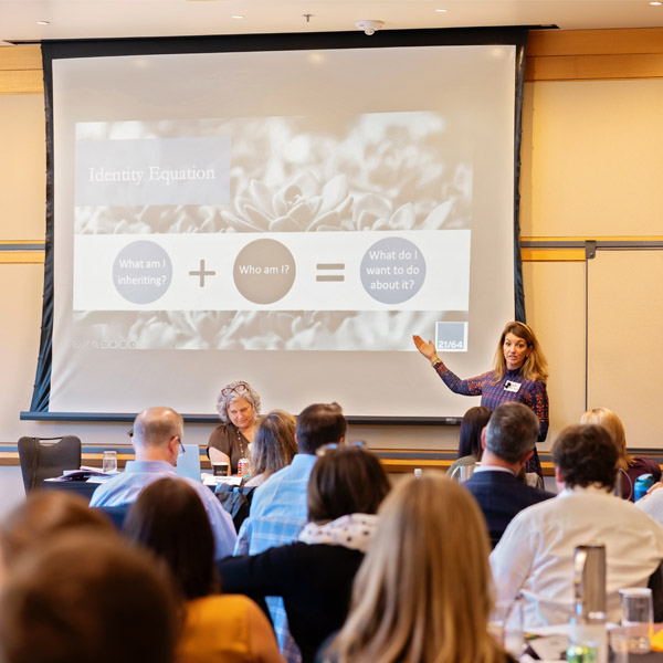 A woman stands and gestures toward a presentation slide on a projector screen in front of an audience. The slide reads “Identity Equation” and features the formula: “What am I inheriting? + Who am I? = What do I want to do about it?”.
