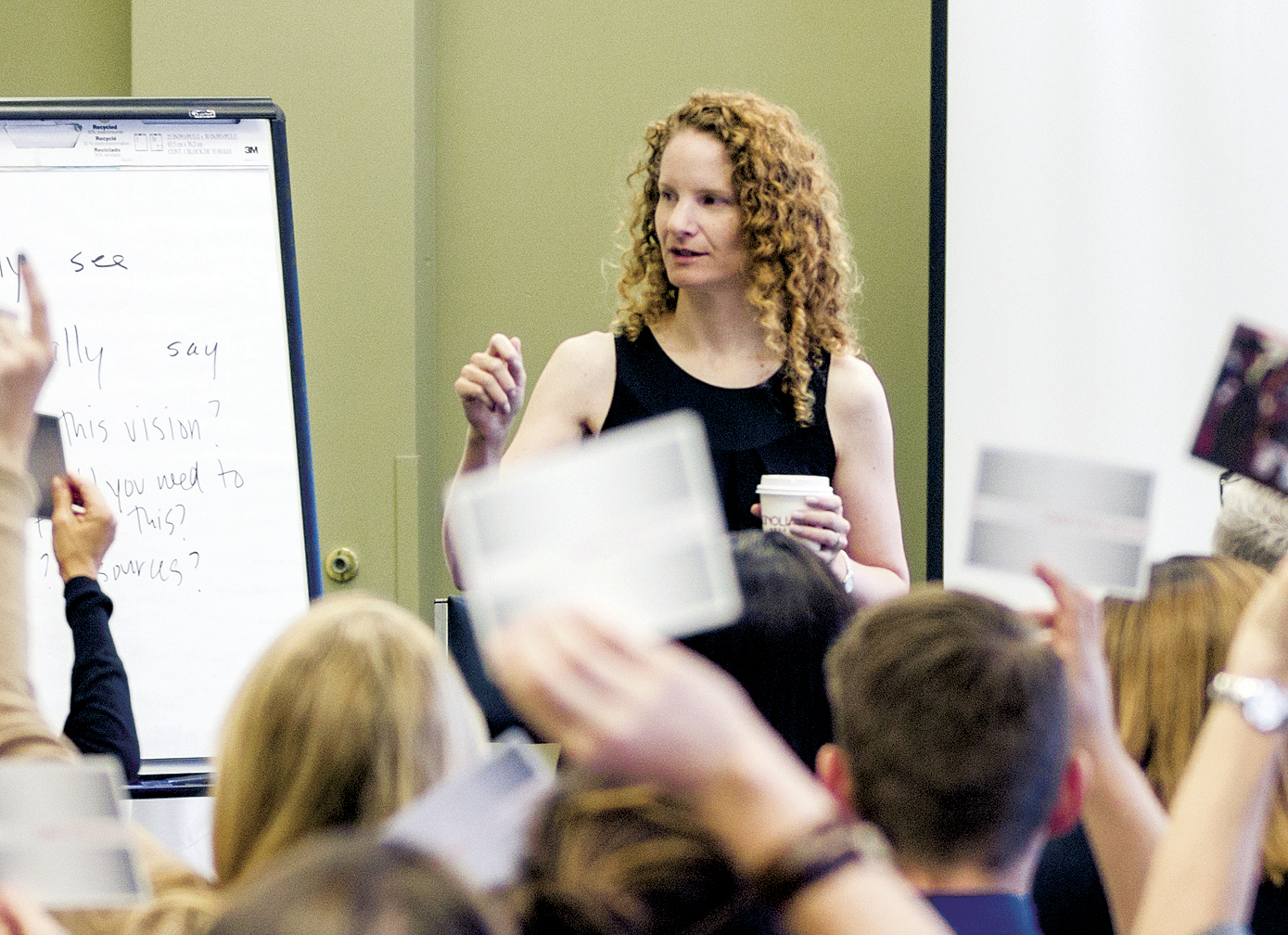 A woman with curly hair stands by a flip chart, holding a cup and speaking to an audience. Several people in the audience raise cards, and the woman gestures as she addresses them.