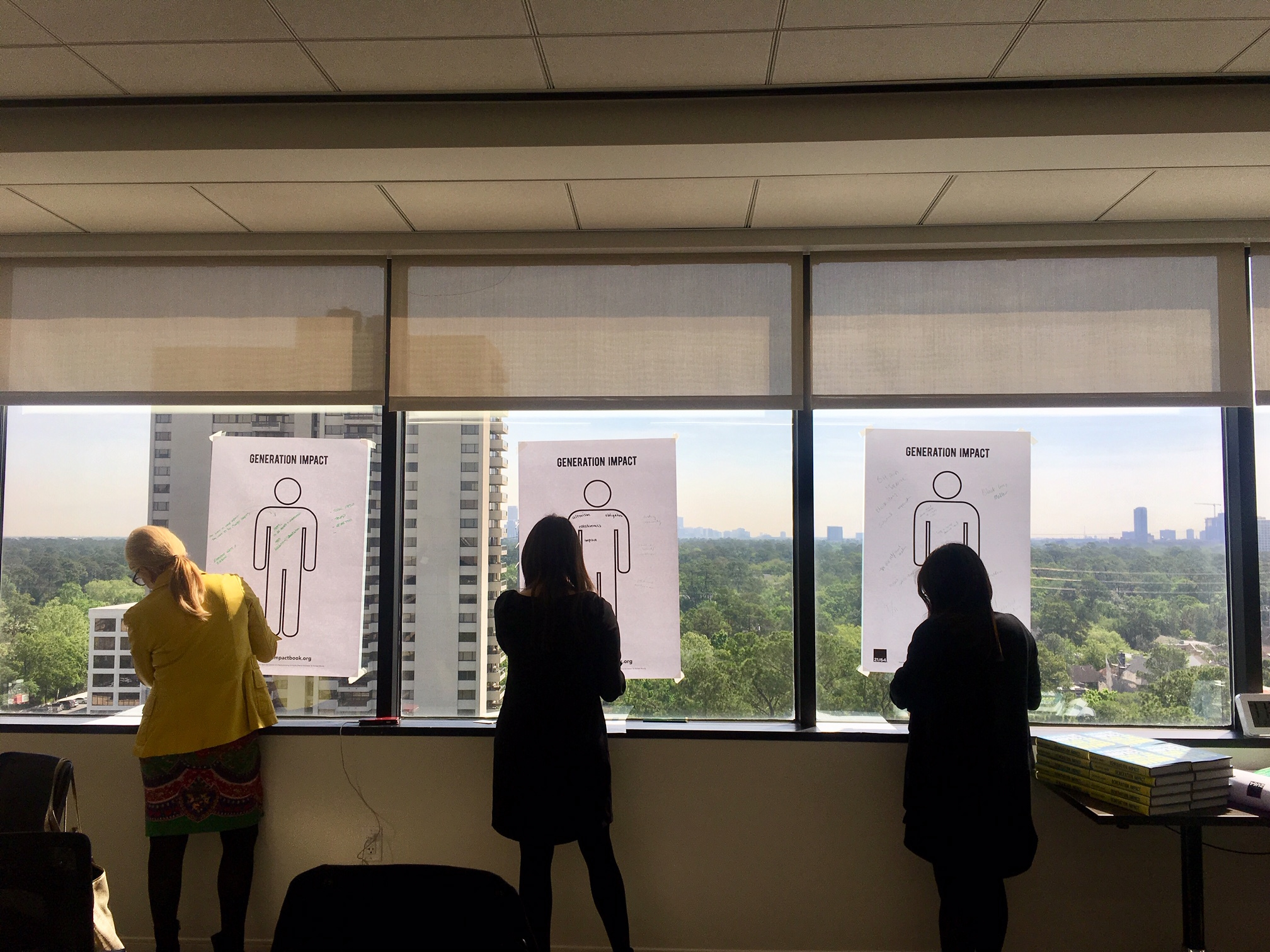 Three people stand by large windows in an office, drawing on posters with human figures labeled GENERATION IMPACT. Tall buildings and trees are visible outside through the windows.