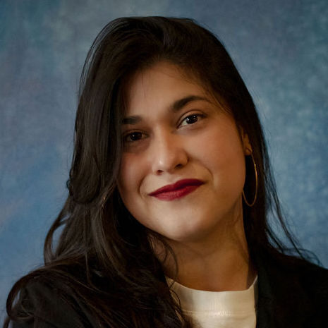 A woman with long dark hair and hoop earrings smiles softly at the camera. She is wearing a dark jacket over a light top, with a blue textured background behind her.
