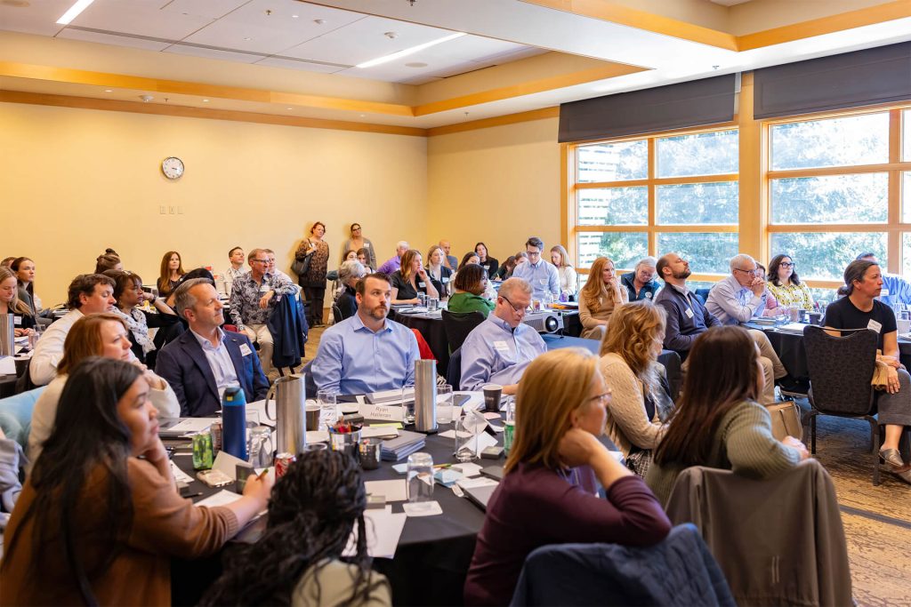 A group of adults sits around tables in a bright conference room, attentively listening to a presentation. Some take notes, while others watch the speaker. Large windows let in natural light.