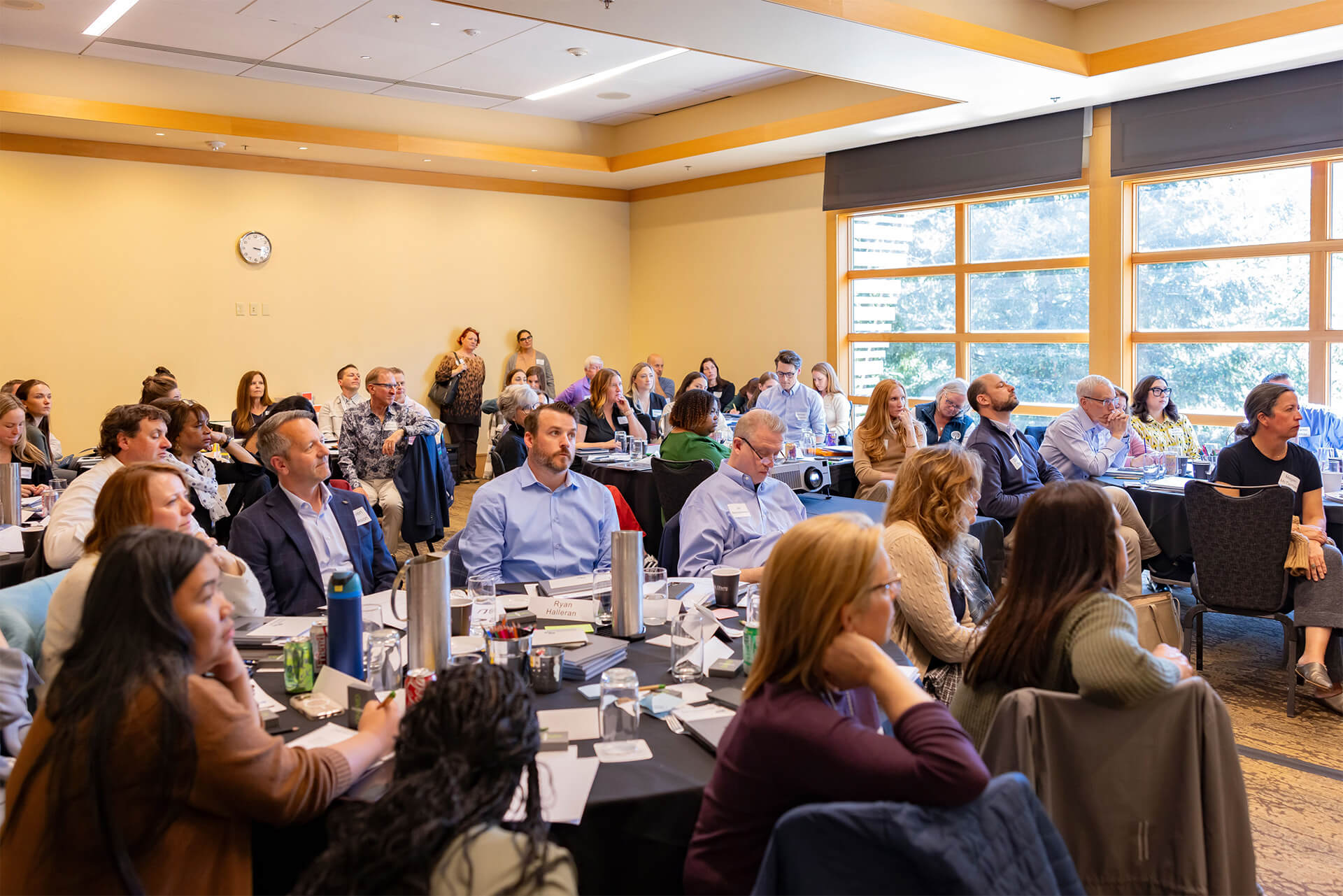 A group of adults sits around tables in a bright conference room, attentively listening to a presentation. Some take notes, while others watch the speaker. Large windows let in natural light.
