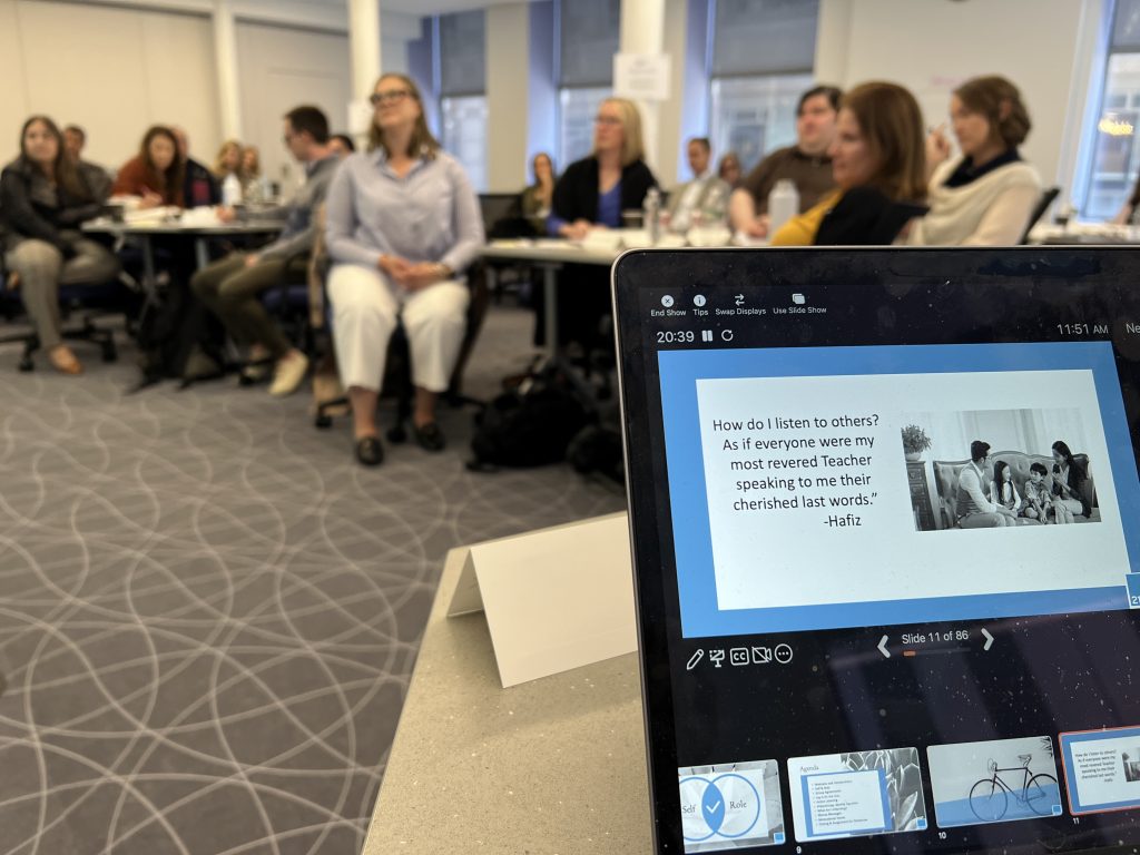 A classroom of adults listens to a presentation. In the foreground, a laptop displays a slide with a quote by Hafiz about listening, alongside a black-and-white photo. The audience appears attentive and engaged.