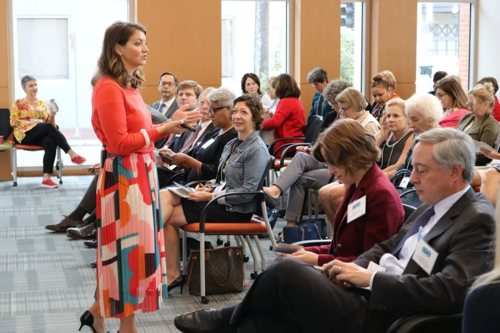A woman in a colorful skirt stands and speaks to a seated audience in a conference room. Attendees, both men and women, listen attentively, some taking notes. The room has large windows and modern office chairs.