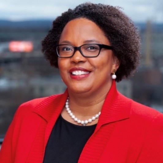 A smiling woman with short curly hair, glasses, and pearl jewelry is wearing a red blazer over a black top. She is standing outdoors with a blurred cityscape in the background.