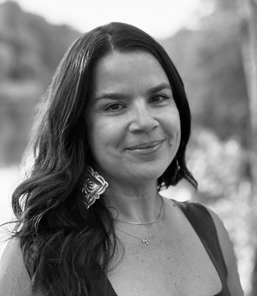A woman with long dark hair, wearing a sleeveless top, a large earring, and layered necklaces, smiles gently while standing outdoors. The background is blurred, emphasizing her face. The image is in black and white.