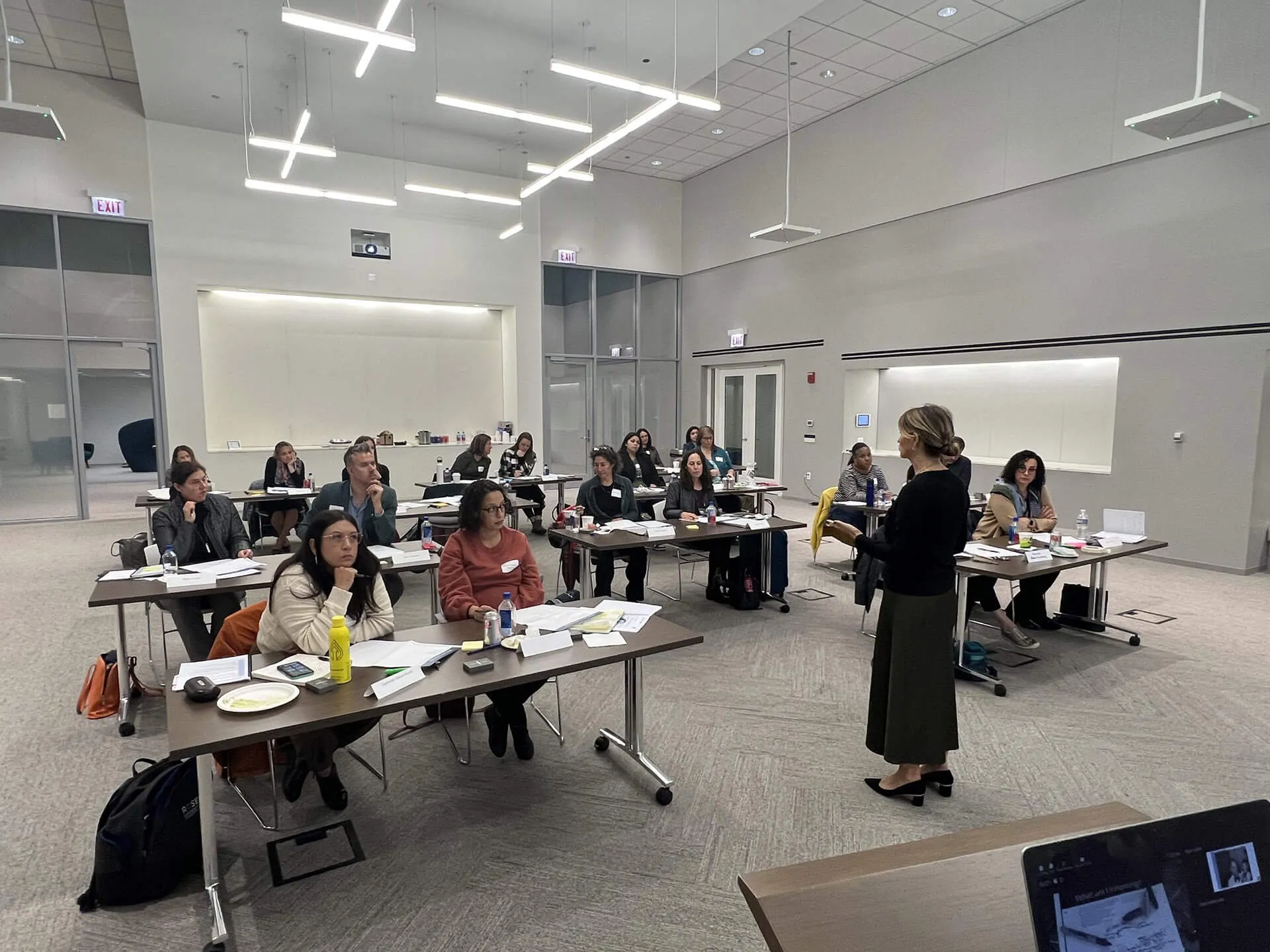 A woman stands and speaks to a group of people seated at tables in a modern, well-lit conference room. Attendees listen attentively, and papers, drinks, and laptops are on the tables.