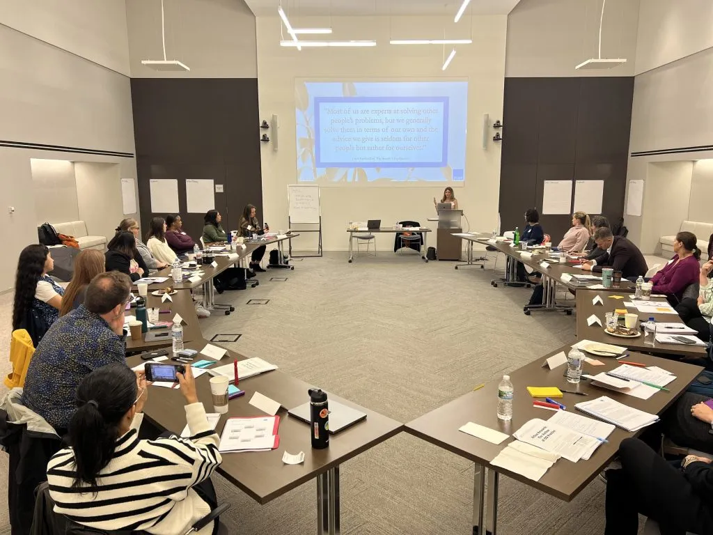A group of people sit around a U-shaped arrangement of tables in a conference room, listening to a presenter at the front. A presentation is projected on the wall behind the presenter. Papers and drinks are on the tables.