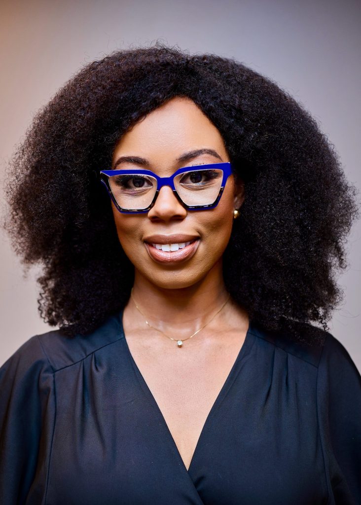 A woman with natural curly hair wears blue cat-eye glasses, a black blouse, and a delicate necklace. She is smiling and stands against a neutral background.