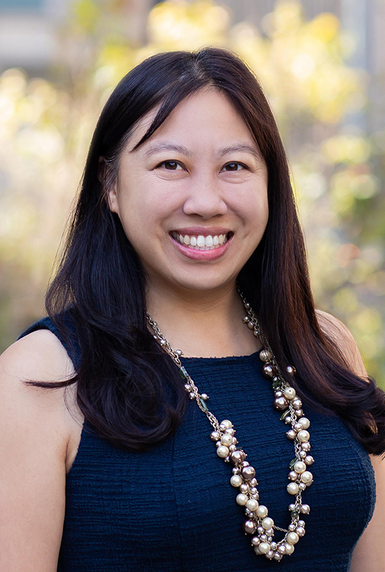 Woman with long dark hair wearing a sleeveless navy dress and a pearl necklace, smiling outdoors with a blurred background of greenery and sunlight.
