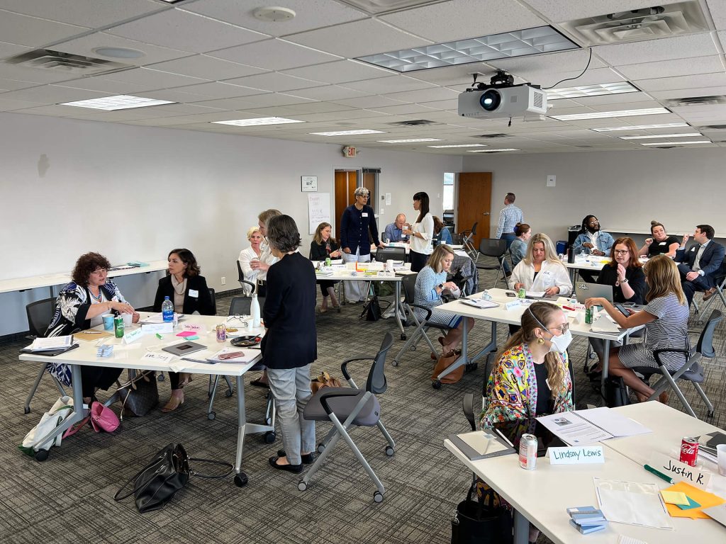 People sit and talk in small groups at tables in a bright conference room with gray carpet and white walls. Papers, drinks, and name cards are on the tables, and a projector hangs from the ceiling.