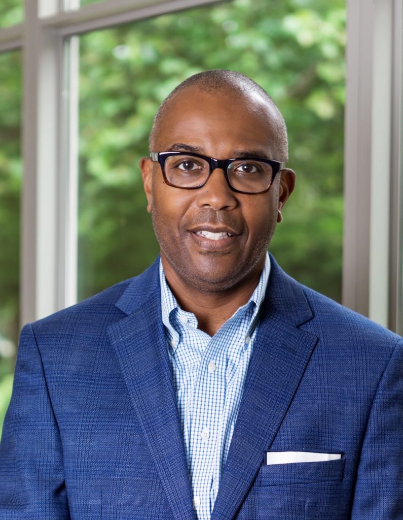 A man wearing glasses and a blue checked shirt with a blue blazer stands in front of large windows with green trees visible outside. He is smiling slightly and a white pocket square is visible in his jacket.