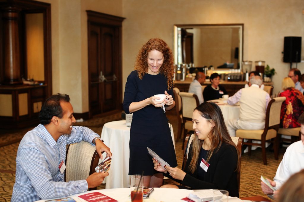 Three people interact around a table at a formal event. A woman stands, smiling, holding papers, while a man and a woman sitting at the table look at cards, engaged in conversation. Other attendees are visible in the background.