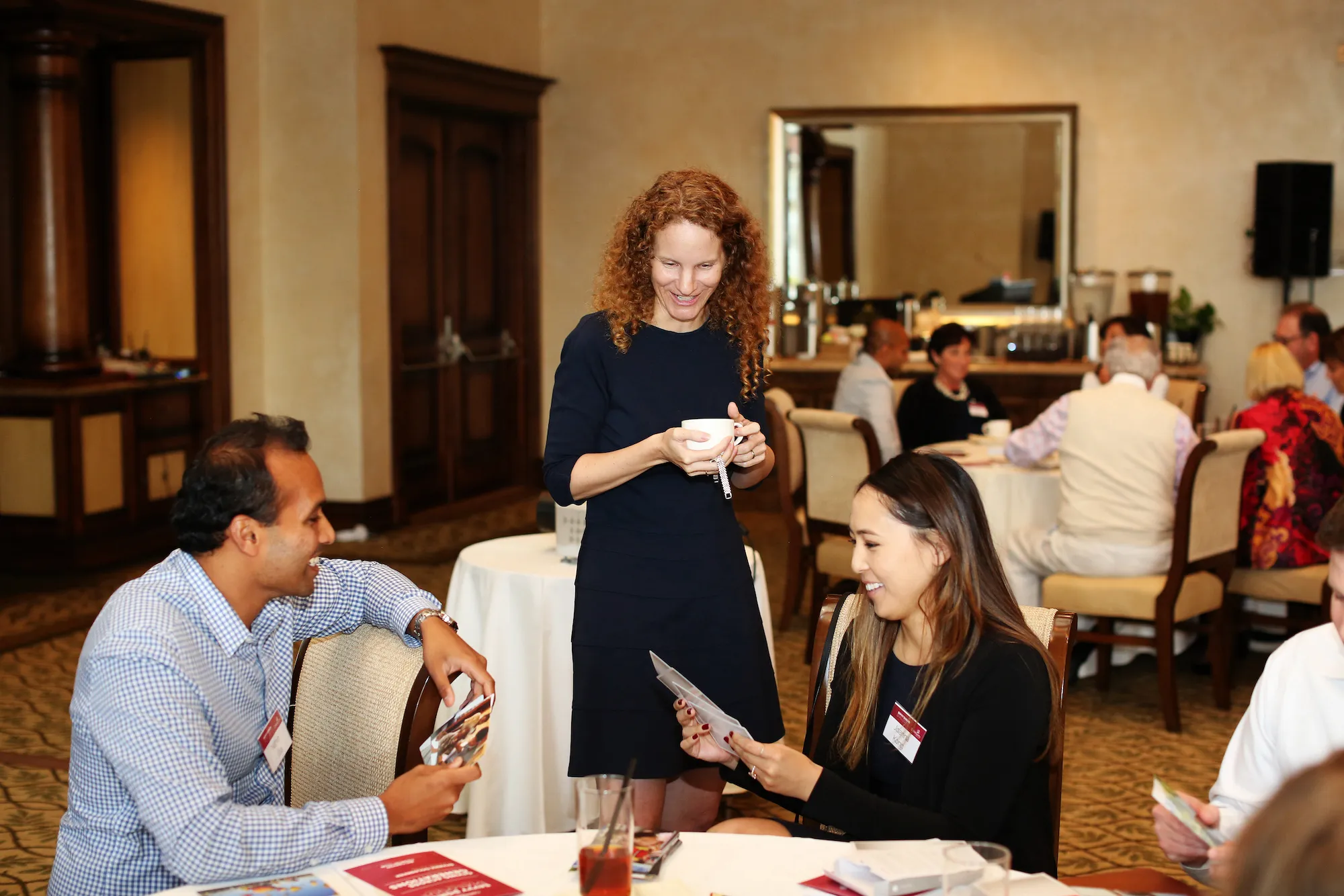 Three people interact around a table at a formal event. A woman stands, smiling, holding papers, while a man and a woman sitting at the table look at cards, engaged in conversation. Other attendees are visible in the background.