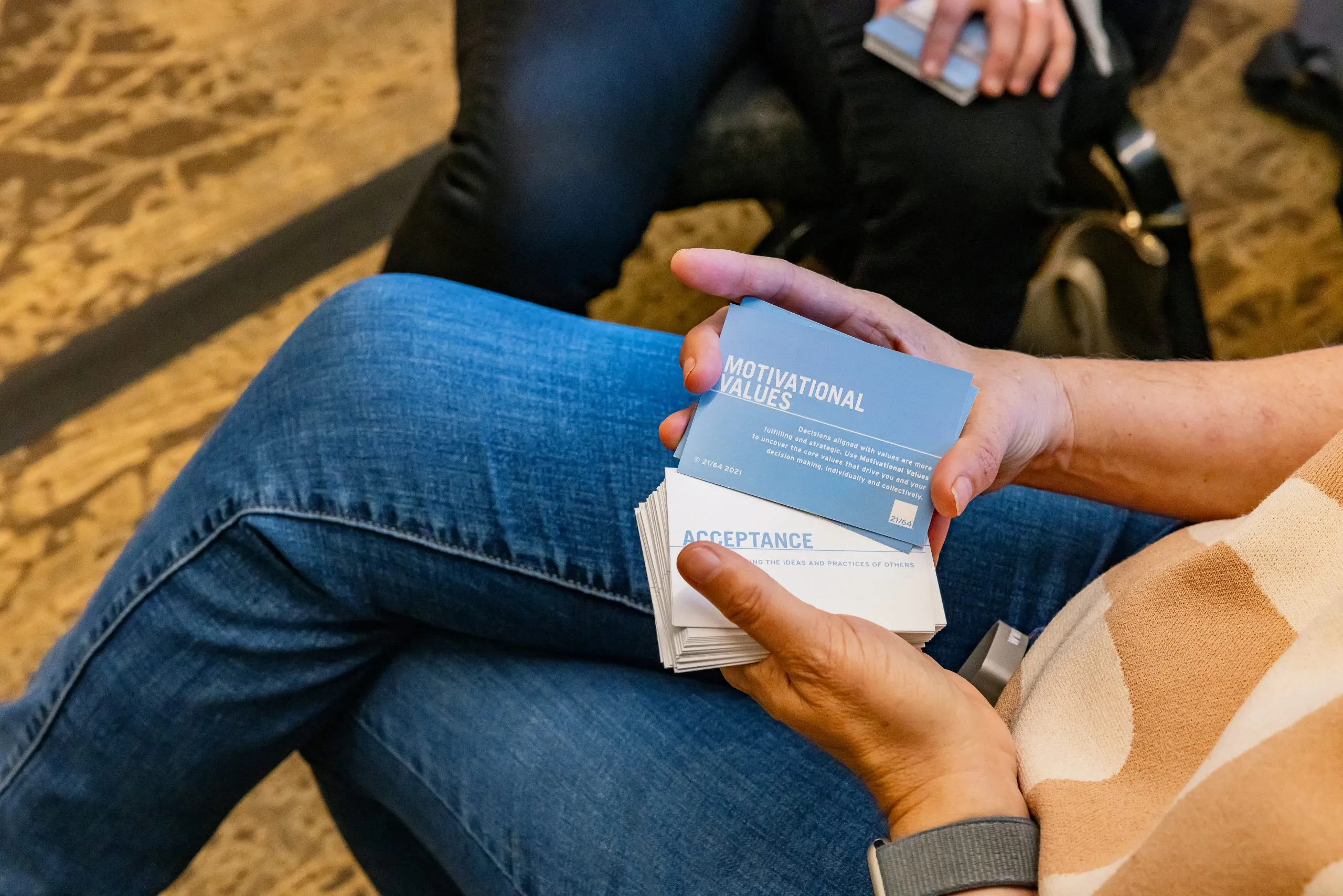 A person in jeans holds a stack of blue cards labeled Motivational Values, with Acceptance visible on the top card; another person sits nearby, partially visible.