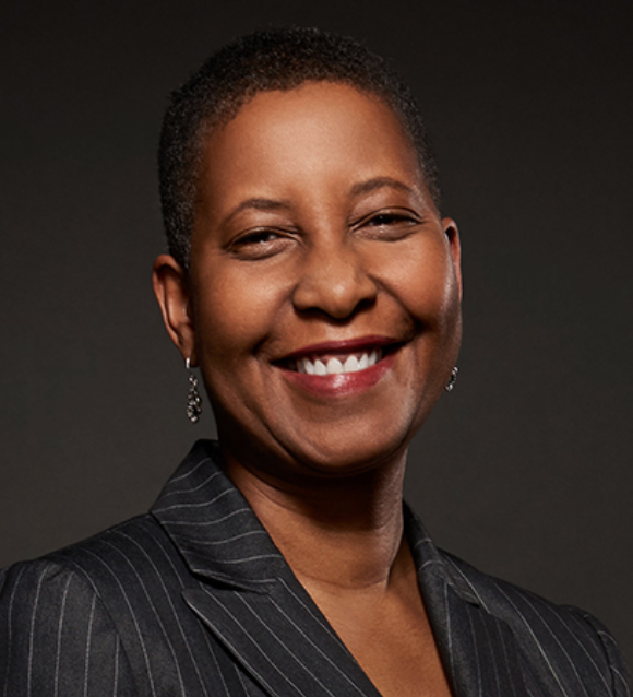 A smiling woman with short hair wearing a pinstripe suit jacket and dangling earrings poses in front of a dark background.