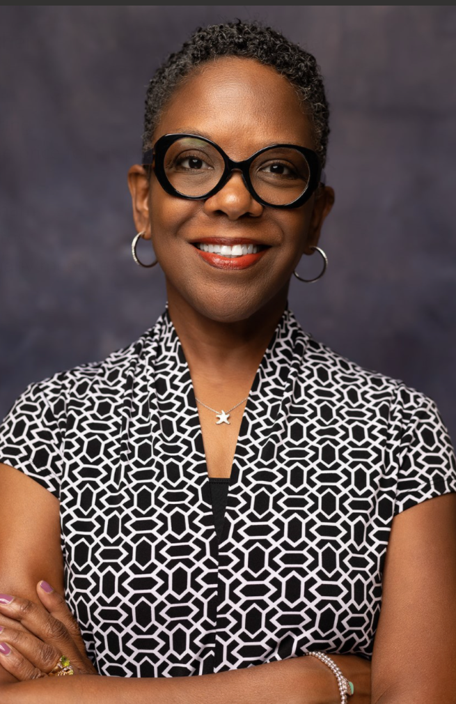 Smiling woman with short curly hair, wearing large round glasses, hoop earrings, and a black-and-white geometric patterned top, stands with arms crossed against a dark studio background.