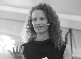 A woman with curly hair, wearing a sleeveless top and a headset microphone, speaks while gesturing with one hand. The photo is in black and white.