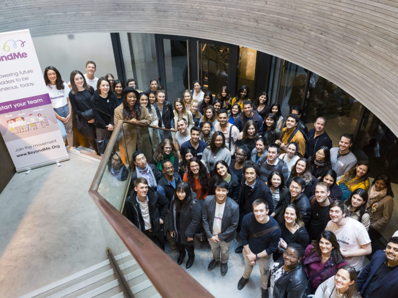 A large, diverse group of people stand smiling on a staircase and surrounding area, posing for a group photo indoors. A banner for BeyondMe is visible on the left. The setting is modern with wood and glass accents.