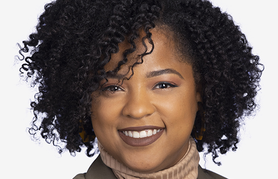 A woman with curly black hair smiles at the camera. She is wearing a beige turtleneck and a dark jacket, with natural makeup and a light-colored background.