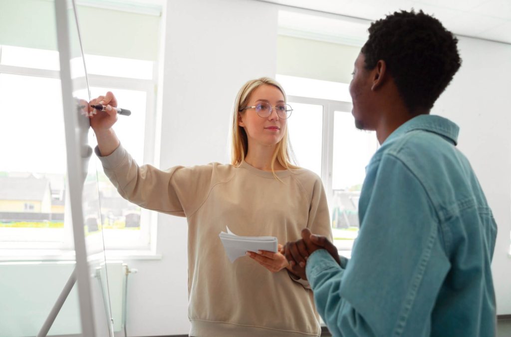Two people are having a discussion in a bright room. One is writing on a whiteboard while holding papers, and the other is facing them, listening attentively.
