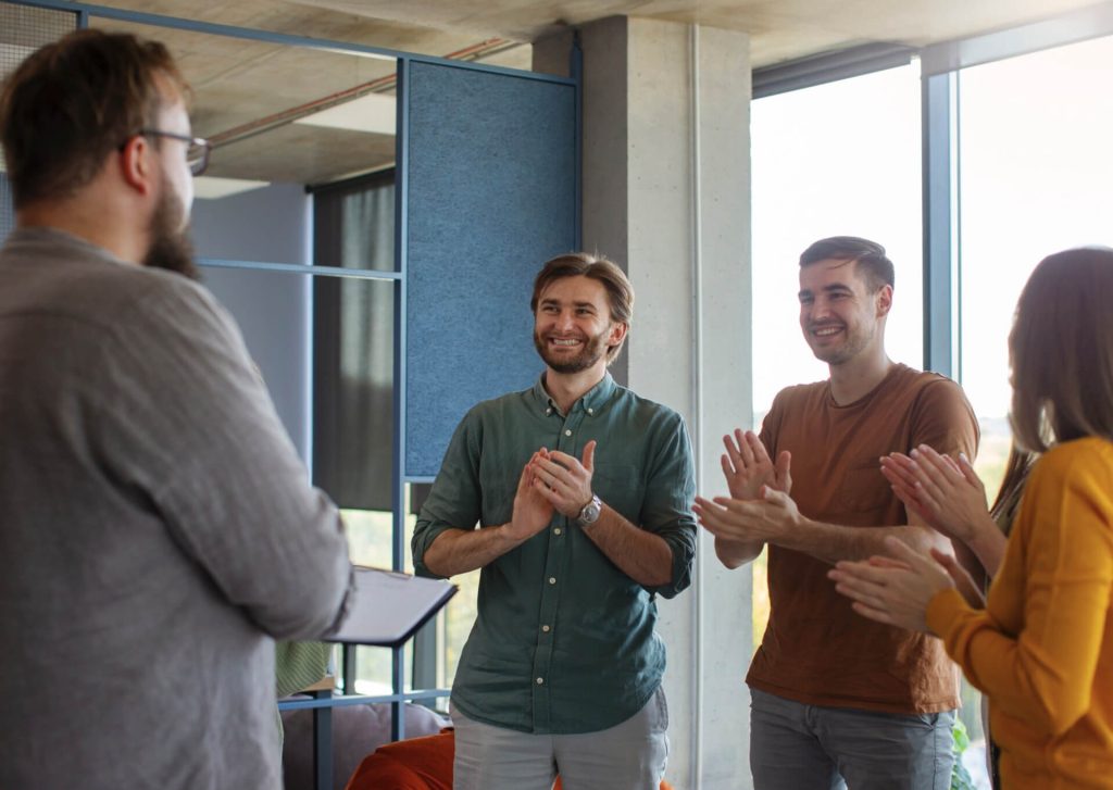 Four people stand in a modern office space. Three of them are clapping and smiling at the person in the foreground, who is holding a clipboard and facing away from the camera. Natural light comes through large windows.