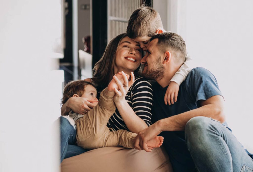 A happy family of four sits closely together at home, smiling and laughing. The mother is in the center, hugging a young child, while the father leans in and an older child embraces both parents affectionately.