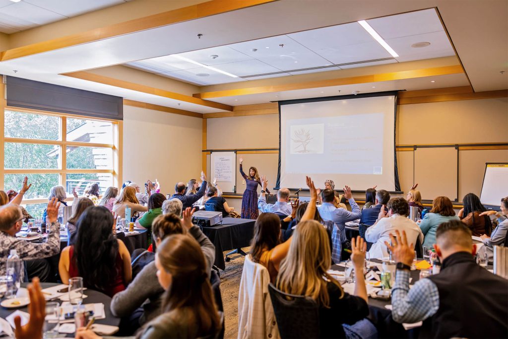 A woman stands at the front of a conference room, speaking to an audience seated at tables; many audience members have their hands raised, and a presentation is displayed on a screen behind her.