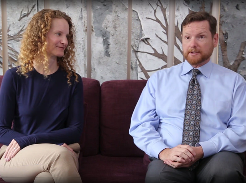 A woman and a man sit on a maroon couch. The woman, with curly hair, wears a navy top and beige pants. The man, with short hair and a beard, wears a blue shirt and patterned tie. They face forward against a nature-themed wall.