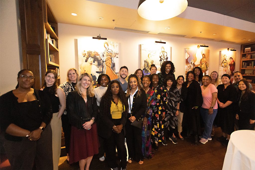 A diverse group of people poses and smiles together in a warmly lit room, standing in front of artwork featuring various figures on the wall behind them.
