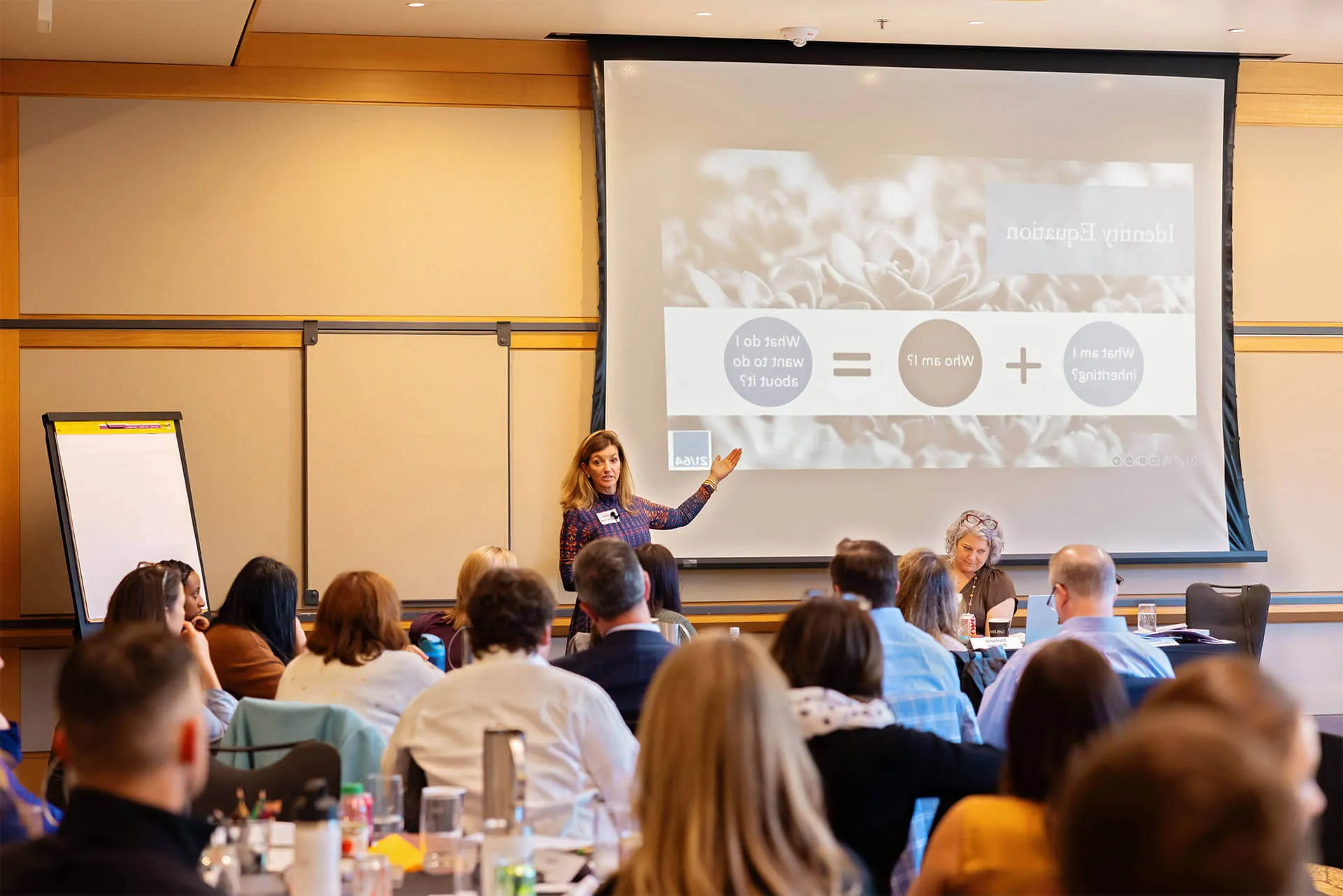 A woman stands and gestures while presenting to a seated group in a conference room. A projection screen behind her displays a diagram. Attendees listen and take notes. A flip chart is visible to the left.