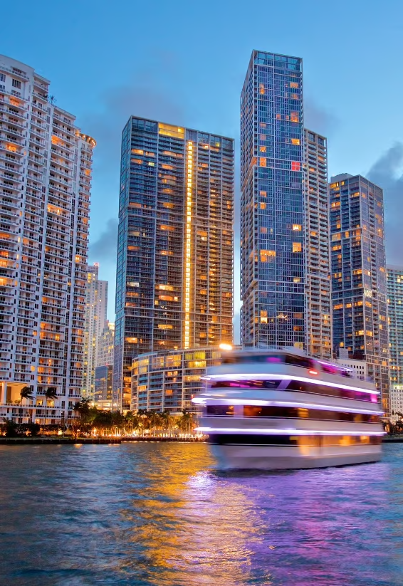 A brightly lit boat moves along the water at dusk, creating a blurred effect. Tall, illuminated skyscrapers tower in the background, reflecting city lights on the water’s surface.