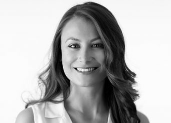 A woman with long, wavy hair smiles at the camera. She is wearing a sleeveless, light-colored top. The background is plain and light, and the image is in black and white.