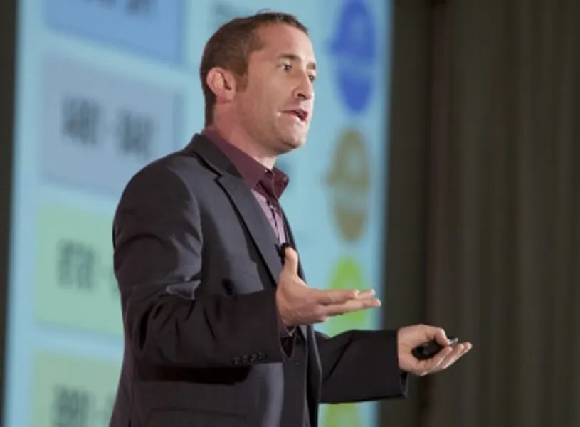 A man in a suit speaks while gesturing with his hands during a presentation, with a colorful projected screen and text visible in the background.