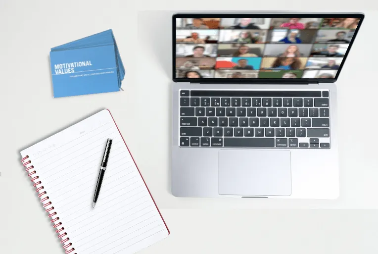 A laptop displaying a video call with many participants, an open notebook with a pen, and a stack of blue cards labeled Motivational Values on a white desk.